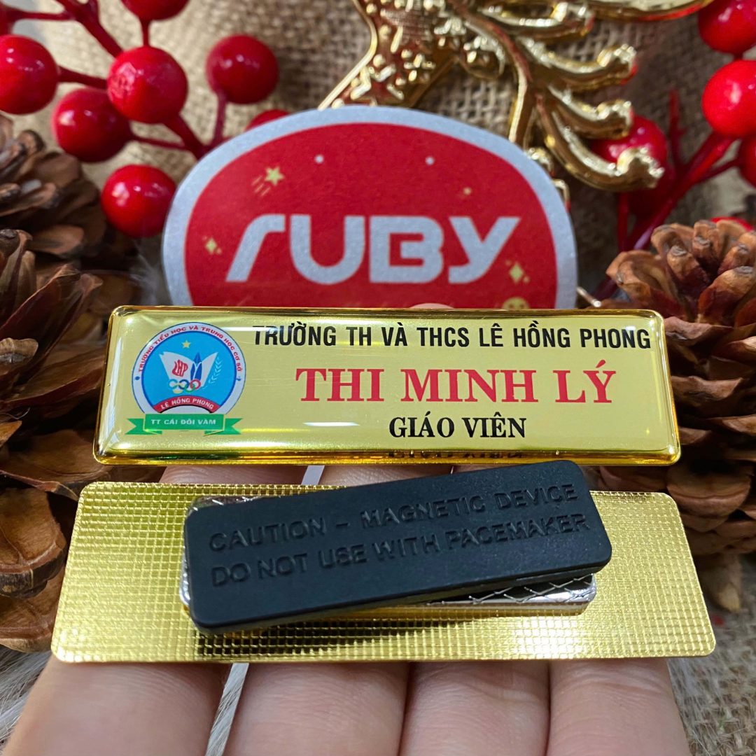 Bảng tên giáo viên nam châm màu vàng của Trường TH & THCS Lê Hồng Phong, in tên “Thi Minh Lý”, kèm mặt sau gắn nam châm không làm thủng áo.